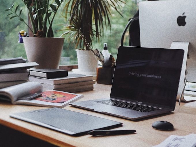 A workspace with a laptop, tablet, notebooks, and potted plants on a wooden desk.