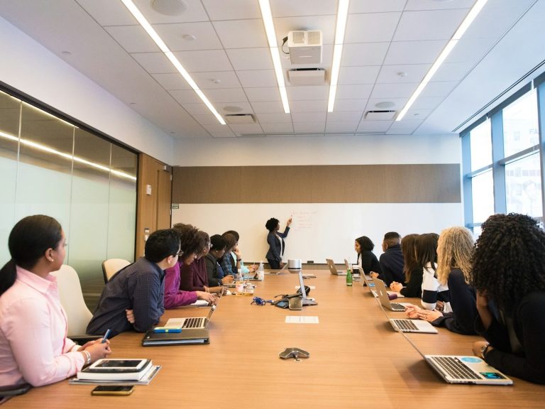 A diverse group of people in a meeting room, listening to a presenter at a whiteboard.