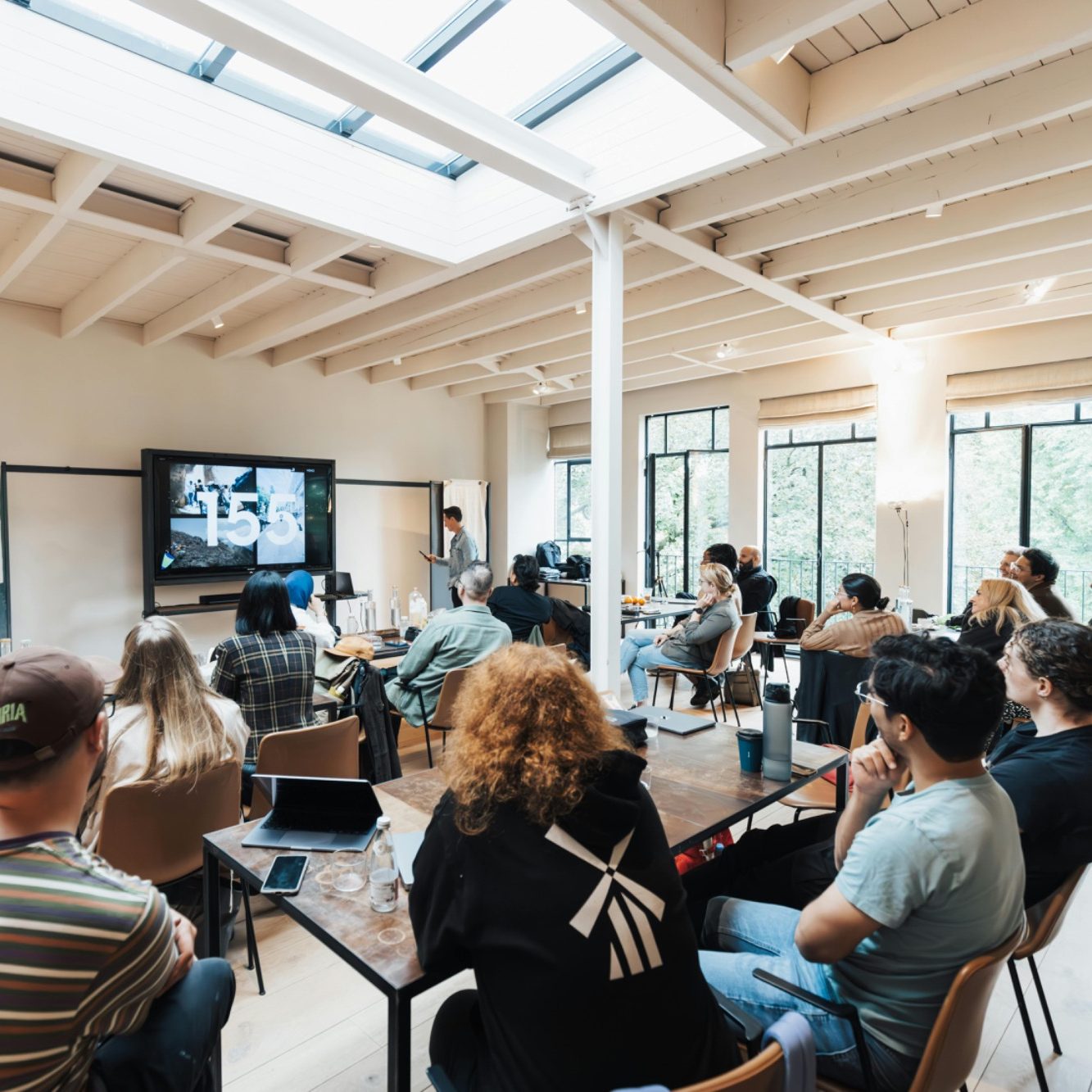 People seated in a bright room, attentively watching a presentation on a screen.